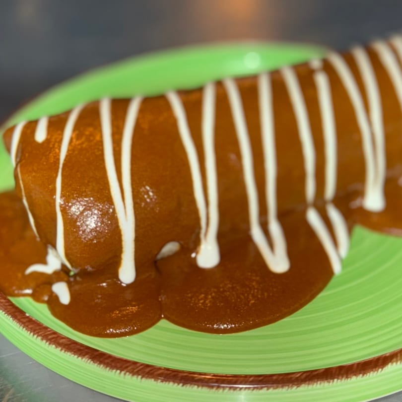 A Mexican burrito covered in rich brown mole sauce and white crema on a green plate.