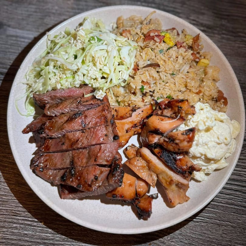 Grilled steak and chicken with fried rice, coleslaw, and macaroni salad on a plate.